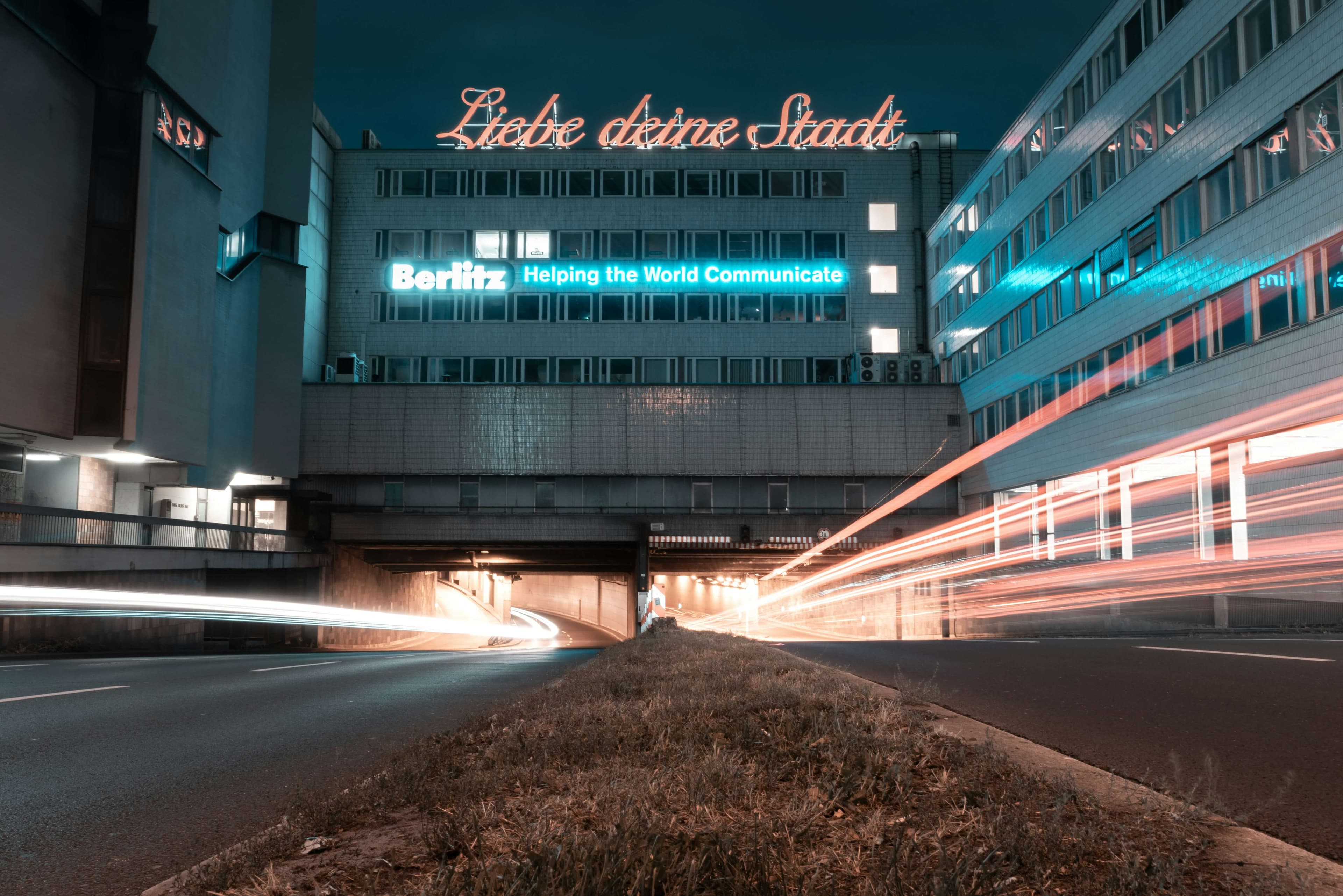 Nächtliche Straßenansicht in Köln mit Lichtspuren von Autos, modernem Bürogebäude und dem leuchtenden Schriftzug 'Liebe deine Stadt' auf dem Dach.