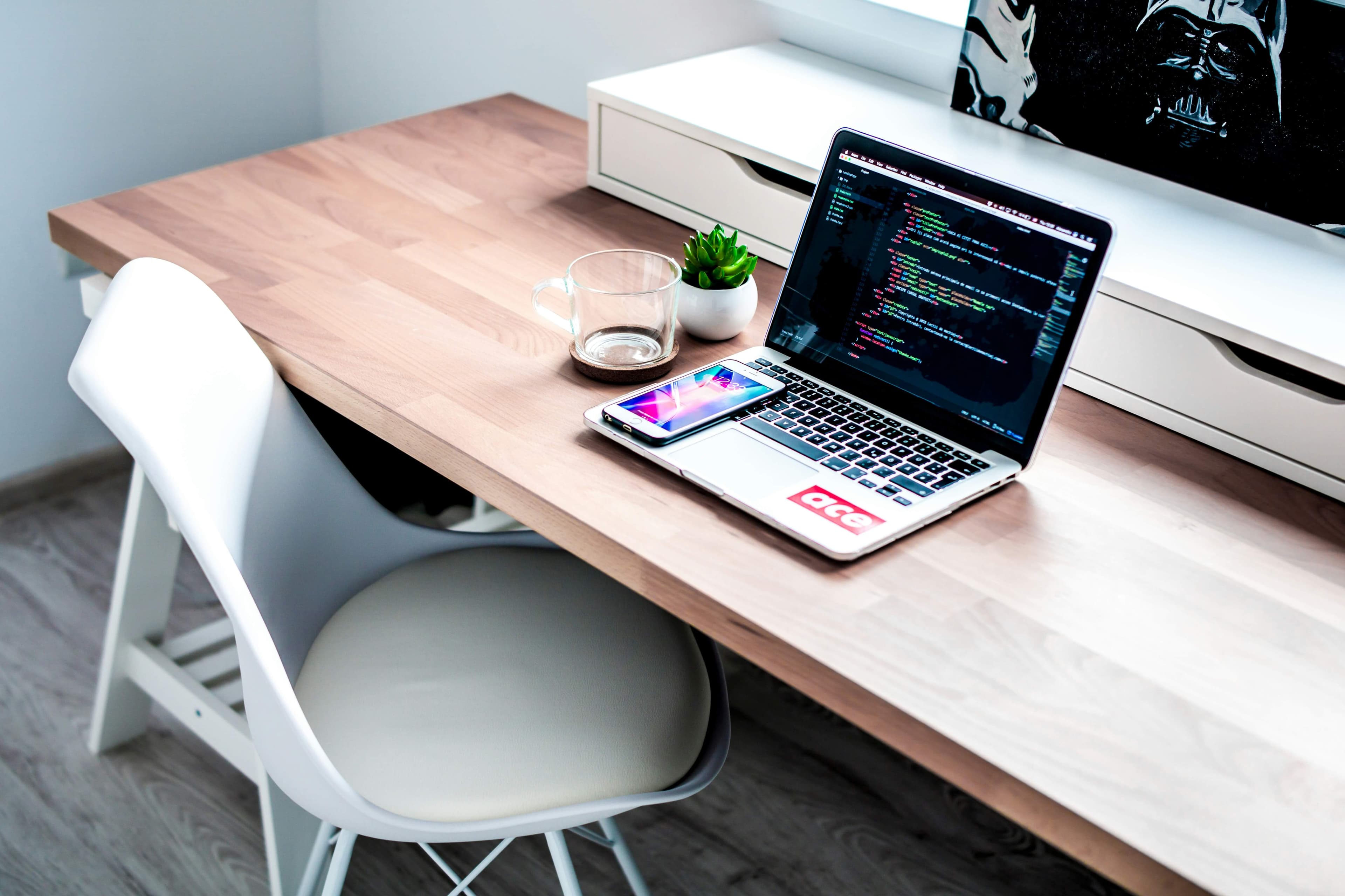 Moderner Arbeitsplatz mit Holzschreibtisch, MacBook mit geöffnetem Code-Editor, Smartphone, leerem Glas, kleiner Pflanze und einem Darth-Vader-Bild im Hintergrund.