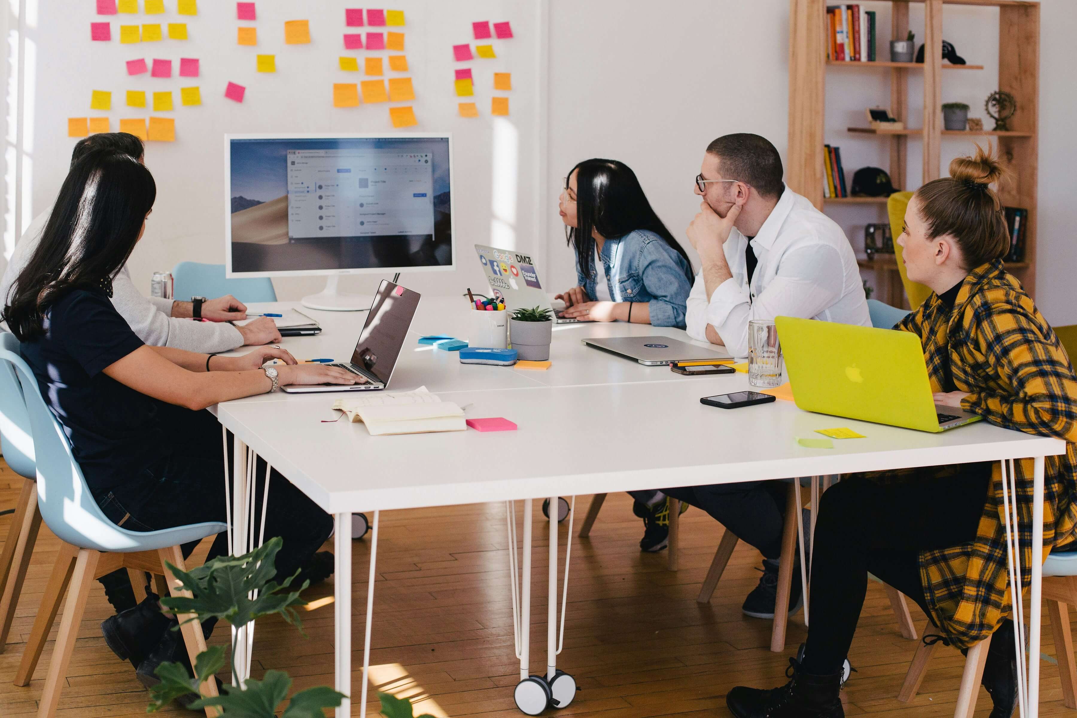 Ein Team von fünf Personen sitzt an einem weißen Konferenztisch in einem modernen Büro und schaut gemeinsam auf einen großen Monitor. Auf dem Tisch stehen Laptops, Notizbücher, Smartphones und bunte Haftnotizen. Im Hintergrund befinden sich viele Post-its an der Wand und ein offenes Regal mit Büchern und Dekoration. Die Atmosphäre wirkt konzentriert und kreativ.